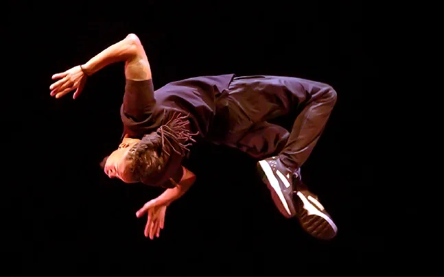 Performer suspended in an acrobatic midair dance move on a dark stage under focused lighting.