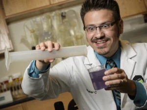 A science presenter in a laboratory coat pours liquid from a large syringe into a clear container filled with purple liquid during a hands-on science demonstration.