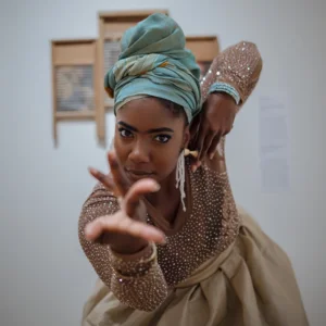 Dynamic portrait of Shanna Woods in a dance pose, extending one hand toward the camera while wearing a headwrap, statement earrings and a sparkling long-sleeve costume in a gallery setting.