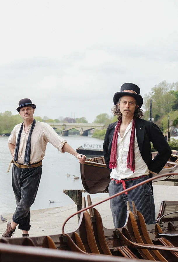 Two musicians from Sons of Town Hall pose beside a wooden boat near the water with acoustic guitars.