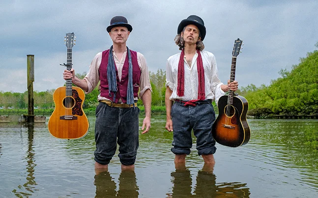 Sons of Town Hall musicians stand in shallow water holding acoustic guitars in a promotional portrait.