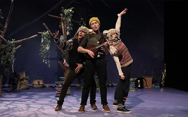 Performers portraying characters from The Boy Who Cried Wolf on stage, including two sheep-inspired costumes and a musician.