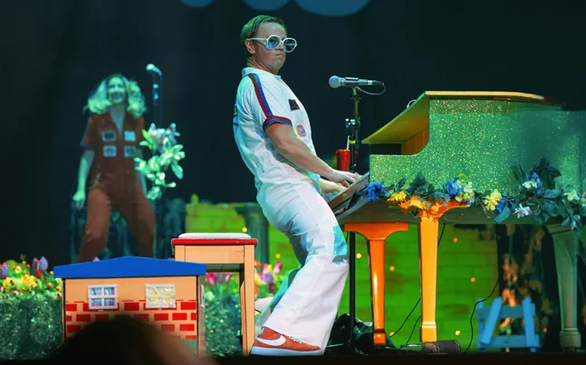 The Rocket Man Show featuring a performer seated at a glittering gold piano on a brightly lit stage. The performer wears a white, retro-inspired costume and oversized glasses while playing piano and singing into a microphone. Colorful floral decorations and theatrical lighting create a vibrant concert atmosphere.