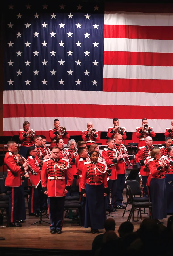 United States Marine Band musicians performing onstage beneath a large American flag. The ensemble wears formal military dress uniforms during a live concert.