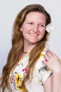 Studio portrait of a person with long wavy hair wearing a white floral-patterned shirt. A white flower is tucked behind one ear, and the person is posed against a light gray background.