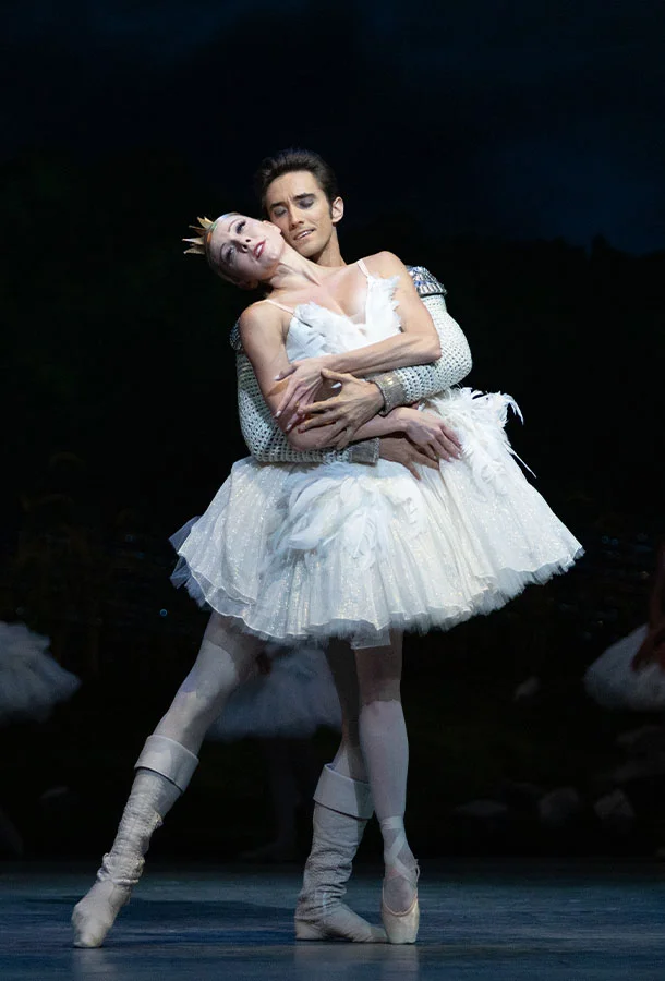 Miami City Ballet's Swan Lake featuring two dancers in a dramatic pas de deux, with the lead performer in a white feathered tutu and pointe shoes. Classical ballet performance showcasing the iconic choreography and timeless elegance of Swan Lake at the Kravis Center.
