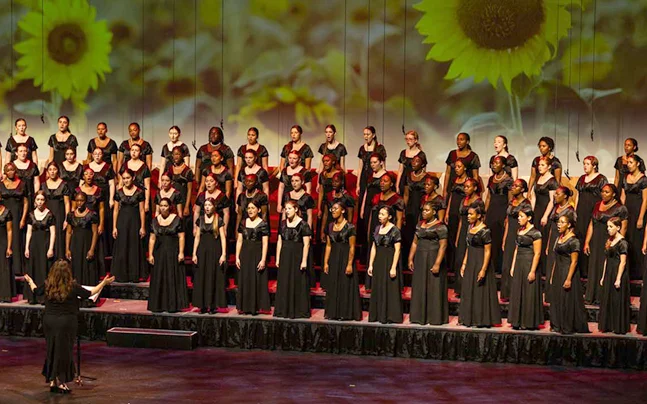 Wide view of a youth choir performing a holiday concert on stage with seasonal lighting and candle imagery projected behind the ensemble.