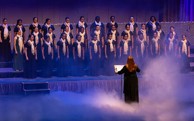 Young performers in formal concert attire sing and gesture in unison during a winter choir performance on stage.
