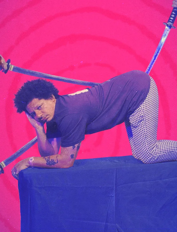 Eric Andre in crouch position on table with swords behind him.